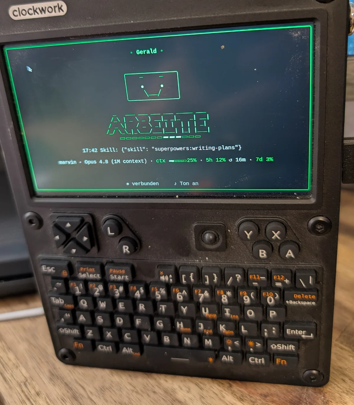 Das uConsole-Handheld auf einem Schreibtisch. Auf dem Display in grüner Schrift: der Name Gerald, ein gezeichnetes Gesicht mit hochgezogenen Brauen, darunter das große Statuswort „ARBEITE“ mit laufendem Balken, der aktuelle Tool-Aufruf und eine Statuszeile mit Modell, Kontextfüllung und Nutzungsgrenzen.